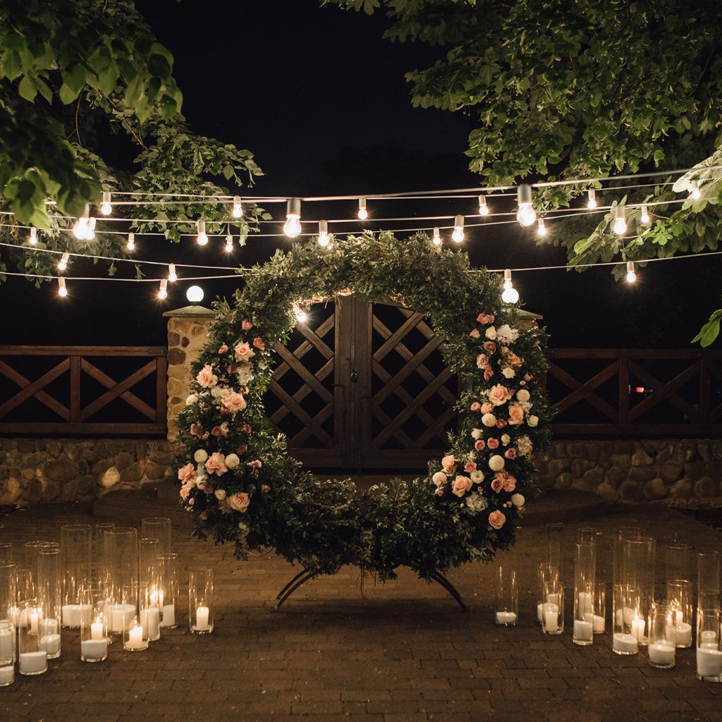 Beautiful photozone with big wreath decorated with greenery and roses in centerpiece, candles on the sides, and garland hanged between trees