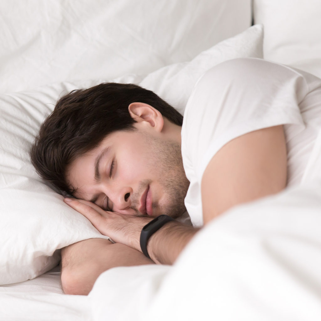 Man is asleep in cozy white bed at night, handsome young guy sleeping at home with wearable electronic device smart watch on his wrist for sleep tracking, monitoring heart rate for healthcare